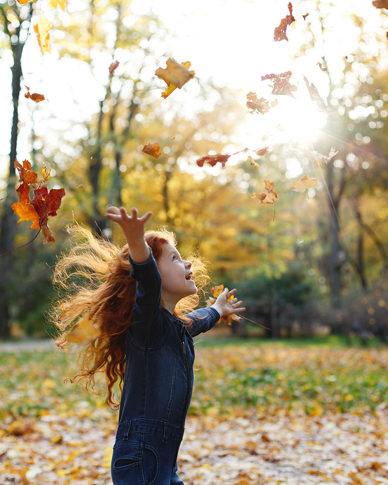 Child is throwing up leaves into the air, she is surrounded by autumn leaves.